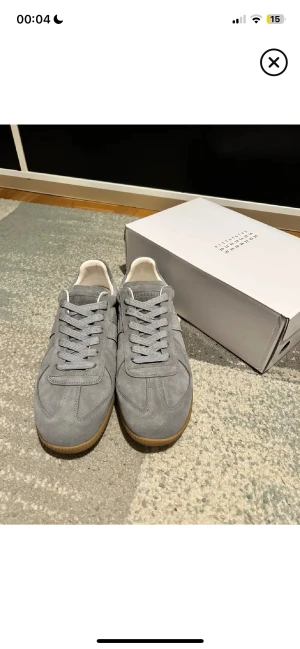 Maison Margiela grå sneakers mocka - Maison Margiela sneakers i grå mocka med klassisk låg siluett och rund tå. Skorna har snörning och en kontrasterande gummisula i beige. Insidan är vit och designen är stilren och minimalistisk, perfekt för dig som gillar exklusiva och cleana sneakers.
