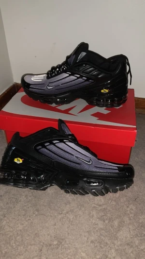 Nike Air Max Plus svart/grå sneakers - Nike Air Max Plus sneakers med svart och grå mesh-överdel, svarta detaljer och reflekterande mönster. Snygg Tn Air-logga på plösen och sidan. Platt sula med synlig Air-enhet och snörning framtill. Perfekt för dig som gillar streetwear och sportig stil.