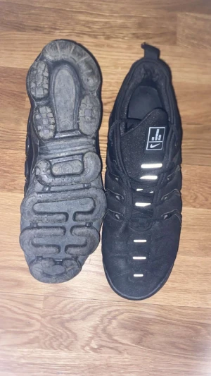 Nike Air Vapormax Plus Svarta 45 - Nike Air Vapormax Plus i helsvart färg med chunky, transparent sula och snygga vågformade detaljer på sidan. Ovandelen är i syntet och mesh med snörning och platt sula. Ikonisk Nike-logga på plösen. Perfekt för dig som gillar streetwear och sportig stil.