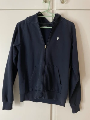 Mörkblå hoodie Peak Performance M - Snygg mörkblå hoodie från Peak Performance i storlek M. Tröjan har huva, dragkedja framtill och två stora fickor. Logga på bröstet och Peak Performance-tryck på ärmen. Perfekt för chill eller träning, mjukt bomullsmaterial.