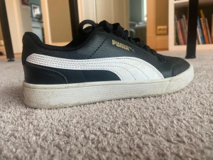 Svarta och vita sneakers från Puma - Snygga svarta sneakers från Puma med vit sula och vit sidodetalj. Skorna har guldfärgat Puma-logo på sidan och hälen, klassisk snörning och rund tå. Ovandelen är i skinn och textil, vilket ger en stilren och sportig vibe.