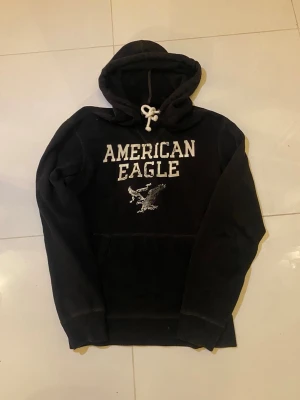 Svart hoodie från American Eagle - Svart hoodie från American Eagle med vit broderad logga och örn på bröstet. Tröjan har huva med vit snörning, klassisk känguruficka och långärmad passform. Väldigt efterfrågat och vintage plagg som är svår att få tag på. Passar även mindre L.