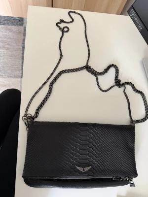 Zadig Voltaire  handväska - Snygg svart axelväska i skinn med reptilpräglad framsida och cool kedjerem. Väskan har dragkedja baktill och ett litet metallmärke framtill. Perfekt storlek för det viktigaste och ger en edgy vibe till din outfit.