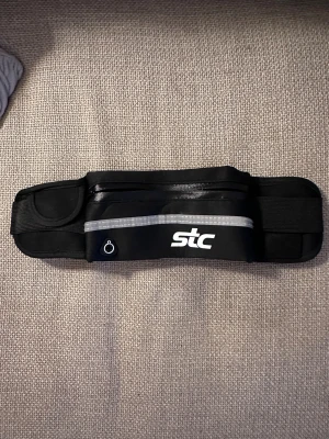 Svart träningsbälte från STC - Säljer ett svart träningsbälte från STC med justerbar rem och smidig plastspänne. Bältet har en praktisk ficka med dragkedja och en liten öppning för hörlurar. Perfekt för att ha mobilen och nycklar nära under löprundan eller gympasset.