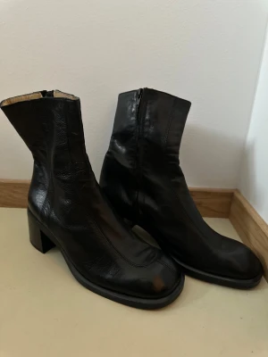 Svarta skinnstövletter - Säljer ett par nya svarta stövletter, tillverkade i Italien. De har en fyrkantig tå, låg klack och dragkedja på sidan. Skorna är gjorda i mjukt skinn och har en stilren, minimalistisk look som passar till många outfits. Nypris 929 kr.