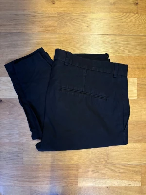Oscar Jacobson Chinos 46 - Svarta chinos från Oscar Jacobson, modell Denz Casual Trousers. Klassisk rak passform med bälteshällor, knapp och dragkedja fram samt diskreta fickor bak. Perfekt för en clean och stilren look. Materialet är mjukt och känns bekvämt mot huden.