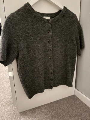 Mörkgrå stickad kofta från H&M - Mysig mörkgrå stickad kofta från H&M med korta ärmar och rund hals. Koftan har knappar hela vägen framtill och en enkel, klassisk design som är lätt att matcha. Helt oanvänd utan prislapp!