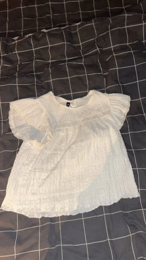 Vit blus med spetsdetaljer från H&M - Säljer en söt vit blus från H&M med korta ärmar och fina spetsdetaljer längs ärmslut och axlar. Blusen är gjord i ett lätt, krinklat bomullstyg och har en luftig passform som är perfekt för varma dagar. Supersnygg till jeans eller kjol!