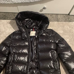 Svart Moncler jacka - Säljer en svart pufferjacka från Moncler med glansig finish och huva. Jackan har dragkedja framtill, två sidofickor och coolt Moncler-band på insidan. Insidan har ett unikt serietidningsmotiv. Perfekt för dig som vill ha en varm och snygg jacka med street vibe. Från NK och köpt för 5000kr