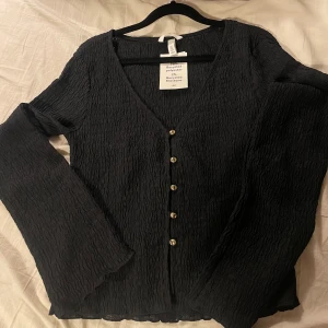 Strukturmönstrad trikptopp  - H&M med v-ringad hals och guldfärgade knappar framtill. Materialet är 98% återvunnen polyester och 2% återvunnen elastan, vilket ger en skön stretchig känsla. Koftan har långärmad design och ett snyggt krinklat tyg som ger extra struktur. Oanvänt endast provat, original pris 179kr