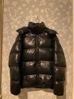 Svart pufferjacka från Everest S men stor i storleken - Säljer en svart, glansig pufferjacka från Everest i storlek S men stor i storleken så jag skulle säga M. Jackan har hög krage, huva och dragkedja framtill. Den är vadderad och riktigt varm, perfekt för kalla dagar. Klassisk och stilren look med diskret logga på ärmen.