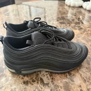 Svarta Nike Air Max 97 sneakers - Säljer ett par helsvarta Nike Air Max 97 sneakers med ikoniska vågformade linjer och synlig luftdämpning i sulan. Skorna har snörning och är tillverkade i en mix av syntet och mesh för en sportig look. Perfekta för dig som gillar streetstyle och komfort.