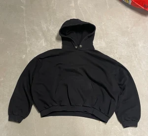 Svart oversized hoodie med huva - Svart hoodie i oversized modell med stor huva och klassisk magficka. Tröjan har ribbade muddar vid ärmslut och nederkant, samt metallöljetter vid snörningshålen. Perfekt för chill dagar och enkel att matcha med allt.