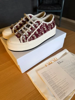 Dior Oblique sneakers vinröd/vit - Snygga Dior Oblique sneakers i vinrött och vitt med ikoniskt mönster och tydlig Christian Dior-logga på plösen. Skorna har vit platt sula, rund tå och klassisk snörning. Ovandelen är i canvas med exklusiv känsla och detaljer i beige. Perfekta för dig som vill sticka ut. Det finns inget kvitto då skorna var en födelsedagspresent för ett år sen. 