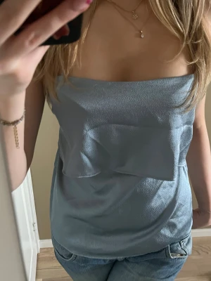 Ljusblå bandeau tubtopp med knyt - Säljer en ljusblå bandeau tubtopp med snygg glansig yta och knytband baktill. Toppen är axelbandslös och har en volangdetalj framtill som ger en cool och trendig look. Perfekt att styla med jeans eller kjol för en fräsch vibe.