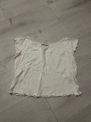 Vit ribbad blus med volangkant - Säljer en söt vit blus från lager 157 med diskret ribbat mönster och volangkant vid ärmar och nederkant. Blusen är kortärmad och har en rund halsringning. Perfekt för dig som gillar en enkel men gullig stil.