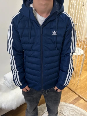 Blå pufferjacka från Adidas med huva - Snygg marinblå pufferjacka från Adidas med klassiska vita ränder längs ärmarna och broderad logga på bröstet. Jackan har huva med snörning, dragkedja framtill och är quiltad för extra värme. Perfekt för kyliga dagar och streetwear-stil.