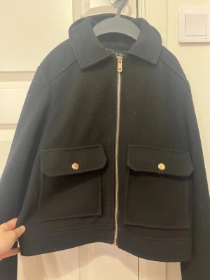 Svart ulljacka med fickor -  svart jacka i ull från ZARA guldiga knappar och dragkedja framtill. Två stora fickor med lock och knappdetaljer Jackan är midjekort och har långa ärmar, perfekt för höst och vinter. säljer för att den inte kommer till användning:(