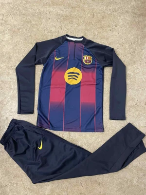 FC Barcelona långärmad fotbollströja Nike - Säljer en långärmad FC Barcelona fotbollströja från Nike med klassiska blå och vinröda ränder, gul Nike-logga och Spotify-sponsor på bröstet. Tröjan har halv dragkedja och är gjord i ett lätt, sportigt material. Matchande mörkblå byxor med gul Nike-logga ingår.