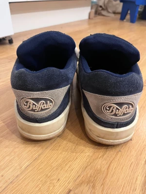 Buffalo sneakers i mocka och blått - Buffalo sneakers med chunky sula i beige och mörkblå mocka. Skorna har rund tå, platt sula och vita snören. Klassisk Buffalo-logga på häl och framsida. Mycket bekväma att gå i och den breda modellen gör att man kan ha tjocka strumpor i. Skulle må bra av en tvätt :)