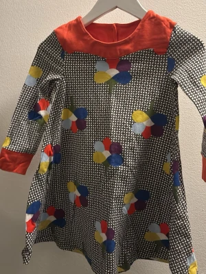 Färgglad blommig klänning med mönster - Unik klänning med svartvitt geometriskt mönster och färgglada blommor i blått, gult, rött och lila. Klänningen har lång ärm, bred röd krage och röda muddar. Perfekt för dig som gillar lekfull stil och vill sticka ut.