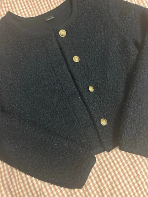 Svart kofta med guldknappar - Säljer en marinblå kofta från Gina Tricot med snygga guldknappar framtill. Koftan är gjord i ett mjukt, bouclé-liknande material och har lång ärm. Perfekt för dig som vill ha en stilren men ändå trendig look.