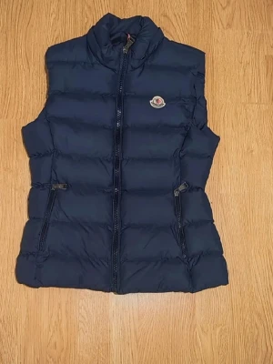  Moncler väst - Säljer en mörkblå dunväst från Moncler med hög krage och två dragkedjeförsedda fickor. Västen har klassisk Moncler-logga på bröstet och quiltad design. Perfekt för lager-på-lager och snygg till både hoodie och t-shirt. Passar någon som är 152cm 