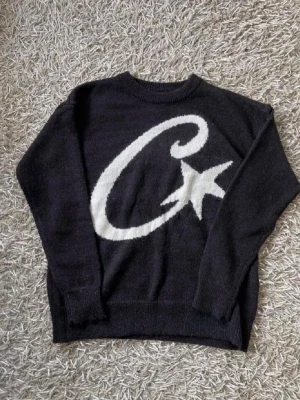 Svart corteiz tröja och mössa - Svart stickad tröja från Corteiz med stor vit stjärna och C på framsidan. Tröjan har rund halsringning och ribbade muddar. Perfekt för dig som gillar streetwear och vill sticka ut med en snygg grafisk design.