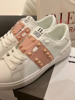 Valentino Rockstud sneakers vit/rosa - Snygga Valentino Rockstud sneakers i vitt skinn med ljusrosa band och ikoniska nitar. Klassisk rund tå, vita snören och coola detaljer på sulan. Perfekta för dig som vill ha en lyxig och trendig look med edge.