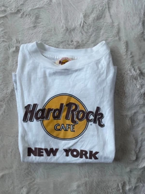 Vit Hard Rock Cafe New York t-shirt - Klassisk vit t-shirt från Hard Rock Cafe New York med ikoniskt tryck i gult och brunt framtill. Rund halsringning och rak passform. Perfekt för dig som gillar retro vibes