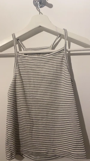 Randig ribbad linnetopp från H&M - Säljer en vit och marinblå ribbad linnetopp från H&M Basic. Toppen har tunna axelband och ett rakt snitt, perfekt för varma dagar. Tillverkad i ekologisk bomull och har ett klassiskt randigt mönster som är lätt att matcha med allt.