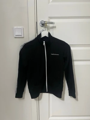  zip Peak Performance kofta  - Svart ziptröja från Peak Performance med vita detaljer och logga på bröstet. Tröjan har hel dragkedja framtill, ribbade muddar och vita ränder med logga längs ärmarna. Perfekt till träning eller chill. Tillverkad i polyester.
