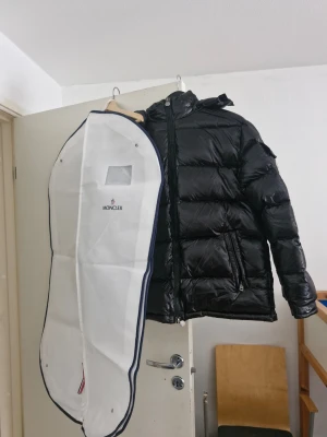 Svart Moncler pufferjacka med huva - Säljer en svart pufferjacka från Moncler med huva och dragkedja framtill. Jackan har två snedställda fickor med dragkedja och Moncler-logga på ärmen. Materialet är glansigt och quiltat, perfekt för kalla dagar. Orginalpris £890. Köpte j Manchester Oktober 21 2025.