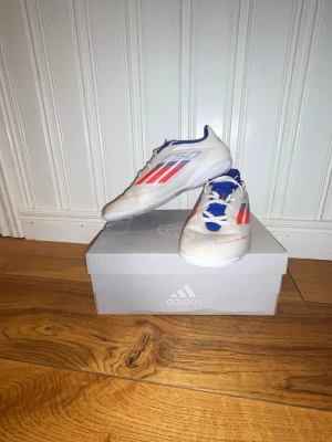 Adidas F50 inomhus/fotbollsskor - Säljer ett par Adidas F50 inomhus/fotbollsskor i vitt med röda och blå detaljer. Skorna har klassisk snörning och är designade för att ge bra grepp på planen. Lätta och smidiga med en sportig look, perfekta för dig som vill sticka ut på matchen.