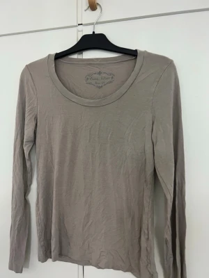 Beige långärmad topp Basic Wear - En enkel och stilren beige långärmad topp från Basic Wear i storlek M. Toppen har rund halsringning och är gjord i ett mjukt bomullsmaterial som känns skönt mot huden. Perfekt att matcha med jeans eller kjol för en clean look.
