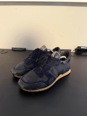 Valentino Garavani Rockrunner sneakers - Snygga Valentino Garavani Rockrunner sneakers i mörkblått med camouflagemönster. Skorna har ovandel i mocka och textil, klassisk snörning och coola nitar på hälen. Sulan är beige med blå detalj och platt sula för skön komfort. Väldigt stor i storleken, jag har 43/44 och de sitter perfekt!
