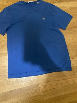 Blå t-shirt från Gant i bomull - Klassisk blå t-shirt från Gant med rund hals och liten broderad logga på bröstet. Tillverkad i mjuk bomull och har en enkel, stilren design som passar till det mesta. Perfekt för dig som gillar basic plagg med snygg passform.  Obs tröjan har ett hål i botten av tröjan knappt synligt 