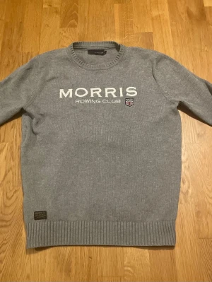 Grå stickad tröja från Morris - Säljer en grå stickad tröja från Morris Rowing Club med broderad logga och text framtill. Tröjan har rund halsringning, ribbade muddar och en klassisk passform. Perfekt för dig som gillar stilren och sportig look.