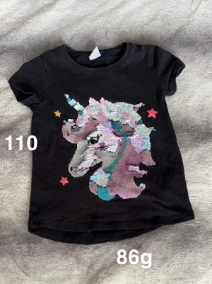 Svart t-shirt med paljett-unicorn - Svart t-shirt med korta ärmar och en stor glittrig enhörning i paljetter på framsidan. Färgglada detaljer i rosa, blått och lila samt små stjärnor runt motivet. Perfekt för dig som gillar magiska motiv och lite extra bling.🌸 Fint använt skick. Storlek 110