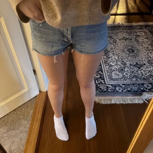 Blå jeansshorts med fransig kant - Säljer ett par klassiska blå jeansshorts med rå, fransig kant och femficksmodell. Shortsen har normal passform och är tillverkade i bomull. Perfekta för varma dagar och ger en avslappnad vibe.