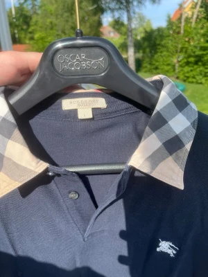 Mörkblå långärmad Burberry piké - Säljer en mörkblå långärmad pikétröja från Burberry med klassisk rutig krage i beige, svart och vitt. Tröjan har knappar vid halsen och Burberrys logga broderad på bröstet. Perfekt för dig som gillar stilrena plagg med en twist.