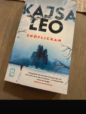 Snöflickan - En spännande bok med vinterkänsla och mystik. Omslaget visar en ensam person som går genom snön mot en skog, vilket ger en känsla av äventyr och hemligheter. Perfekt för dig som gillar gripande berättelser och vill ha något utöver det vanliga.