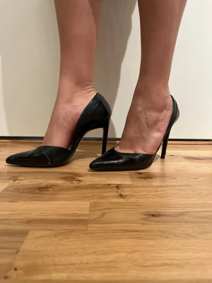 Svarta pumps från Zara Basic - Säljer ett par svarta pumps från Zara Basic med spetsig tå och hög klack. Skorna har en snygg blandning av blankt och strukturerat material som ger en cool kontrast. Perfekta för dig som vill ha en stilren och edgy look.