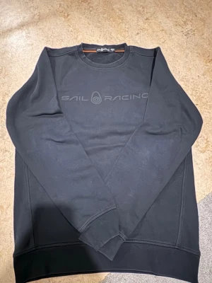 Svart sweatshirt från Sail Racing M - Säljer en svart sweatshirt från Sail Racing i storlek M. Tröjan har rund halsringning, diskret logga framtill och är tillverkad i mjuk bomullsmix. Perfekt för dig som gillar stilren och enkel design.