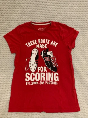 Röd t-shirt med fotbollstryck från Rebel - Röd t-shirt från Rebel med tryck av fotbollsskor och texten 'These boots are made for scoring, eat, sleep...play football' i vitt. Klassisk rund halsringning och korta ärmar. Perfekt för dig som älskar fotboll och vill sticka ut med ett sportigt statement.