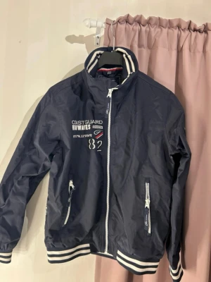 Marinblå vindjacka från Five Seasons - Säljer en marinblå vindjacka från Five Seasons med vita detaljer och broderade loggor på bröstet. Jackan har dragkedja framtill, två fickor med dragkedja och ribbade muddar med vita ränder vid ärmslut och nederkant. Perfekt för blåsiga dagar!
