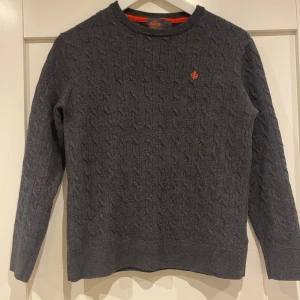 Mörkgrå kabelstickad tröja Morris - Säljer en mörkgrå kabelstickad tröja från Morris i 100% merinoull. Tröjan har rund hals, lång ärm och en orange broderad logga på bröstet. Perfekt för dig som gillar klassisk stil med en twist. Tröjan har krympt och sitter som en xs