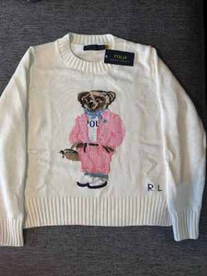 Vit stickad tröja Polo Ralph Lauren - Vit stickad tröja från Polo Ralph Lauren med ikonisk björn i rosa outfit på bröstet. Rund halsringning, ribbade muddar och RL-broderi nedtill. Perfekt för dig som vill ha en klassisk men lekfull look.