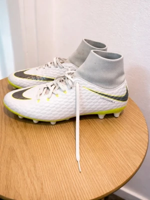 Nike Hypervenom vita fotbollsskor - Säljer ett par Nike Hypervenom fotbollsskor i vitt med grå och neongula detaljer. Skorna har hög strumpliknande krage, snörning och dobbar för spel på gräs. Swoosh-logga på sidan och tå, samt textur på ovandelen för bättre bollkontroll.