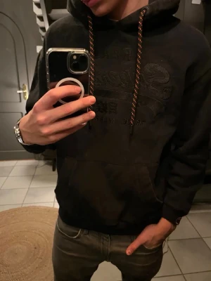Svart superdry hoodie - Säljer en svart superdry hoodie med luva och känguruficka på magen. Han på bilden är 180 och väger 70kg. Storlek S. Hör av dig vid frågor eller liknande🙌